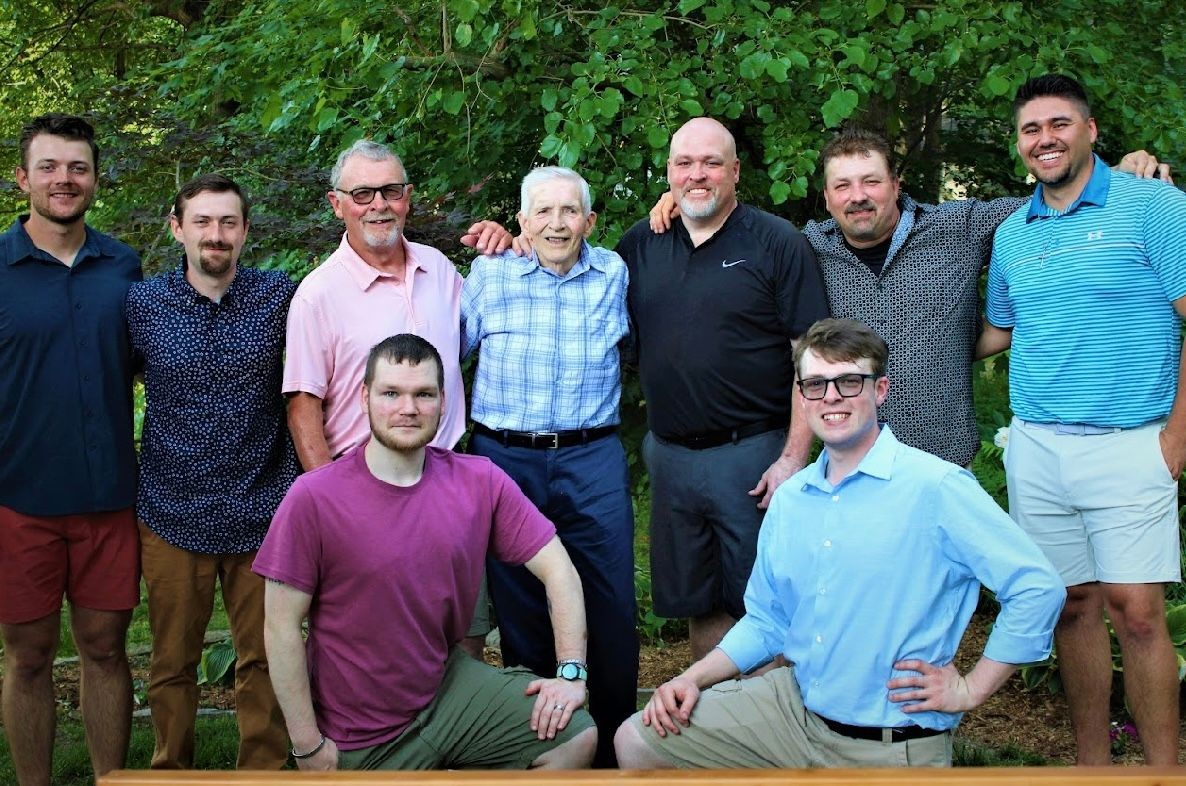 Group photo of several men standing and kneeling outdoors with green foliage in the background.